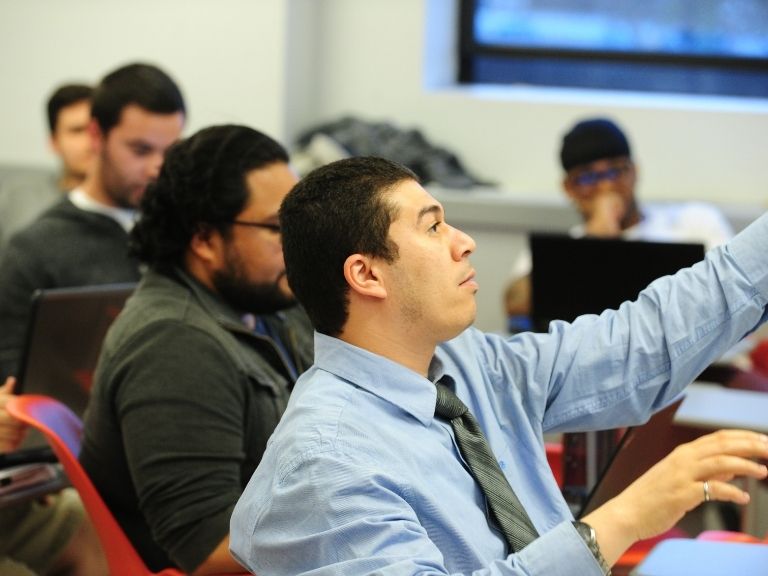 man with dark hair raising his hand in a classroom 